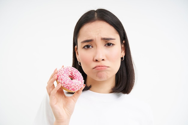 Zdenerwowana szlochająca dziewczyna nie może jeść fast foodów pokazuje pyszne przeszklone pączki chce ugryźć białe tło