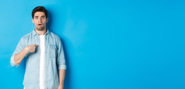 Bezpłatne zdjęcie zdenerwowany mężczyzna wskazujący na siebie i wyglądający na zdezorientowanego stojącego w zwykłych ubraniach na niebieskim tle