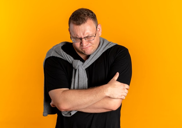 Zdenerwowany mężczyzna z nadwagą w okularach na sobie czarny t-shirt z rękami skrzyżowanymi stojąc na pomarańczowej ścianie