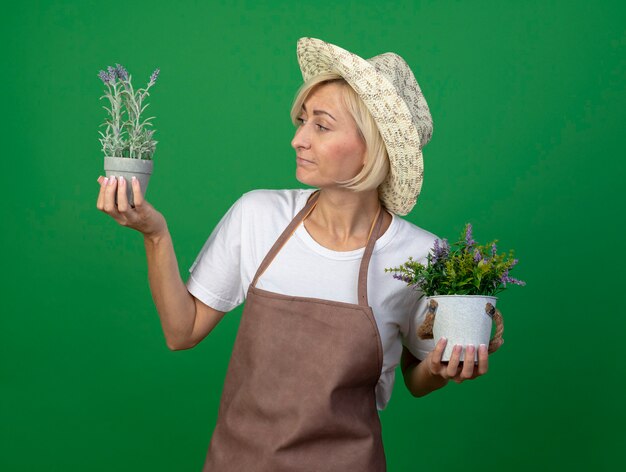 Zdezorientowana blondynka ogrodniczka w średnim wieku w mundurze w kapeluszu trzymająca doniczki patrząca na jedną z nich odizolowaną na zielonej ścianie