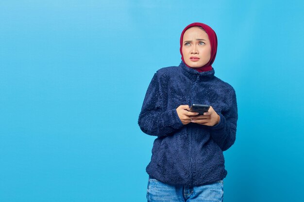 Zdezorientowana młoda azjatka używająca smartfona i patrząca z boku na białym tle na niebieskim tle