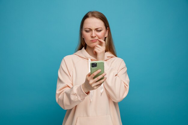 Zdezorientowana młoda blondynka dotyka kącika ust palcem trzymając i patrząc na telefon komórkowy