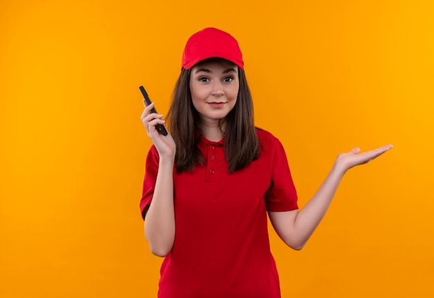 Zdezorientowana młoda kobieta dostawy na sobie czerwoną koszulkę w czerwonej czapce trzyma telefon na odizolowanej pomarańczowej ścianie