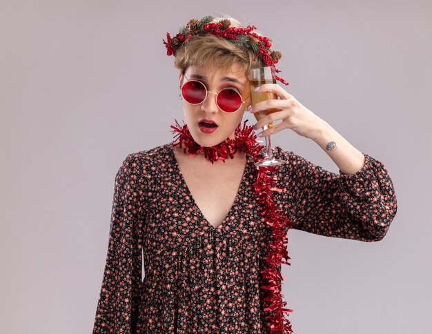 Zdezorientowana młoda ładna dziewczyna ubrana w świąteczny wieniec na głowę i świecącą girlandę wokół szyi w okularach dotykających głowy kieliszkiem szampana, patrząc na kamery na białym tle