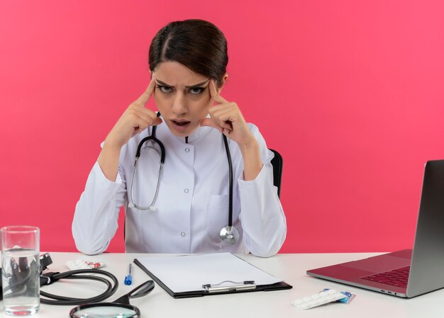 Zdezorientowana młoda lekarka w szlafroku medycznym ze stetoskopem siedząca przy biurku pracuje na komputerze z narzędziami medycznymi kładąc palce na głowie na odizolowanej różowej ścianie z miejscem na kopię