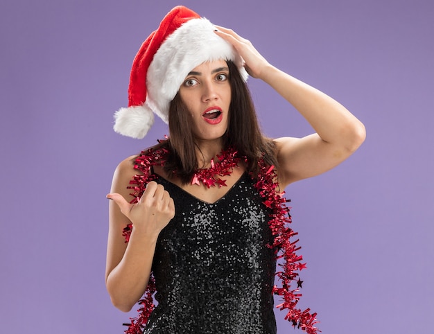 Zdezorientowana młoda piękna dziewczyna ubrana w świąteczny kapelusz z girlandą na szyi po bokach, kładąc rękę na głowie na białym tle na fioletowym tle z miejsca na kopię
