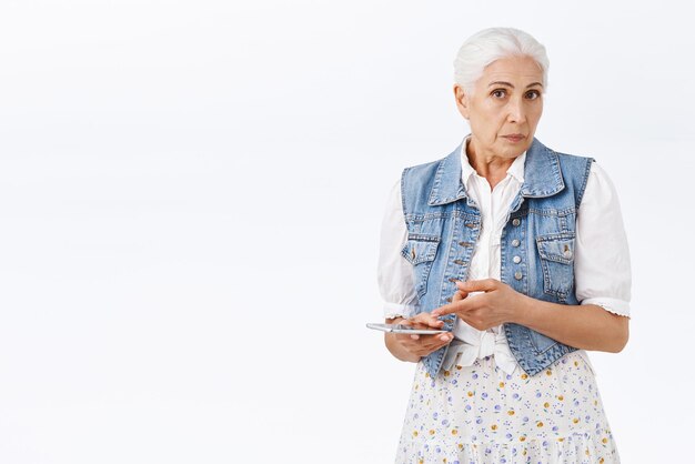 Zdezorientowana, przesłuchiwana starsza kobieta, która nie może zrozumieć, o co chodziło w rozmowie telefonicznej z przytrzymaniem smartfona wskazującego na urządzenie mobilne, gapieniem się na aparat zdziwiony i sfrustrowany stojący na białym tle