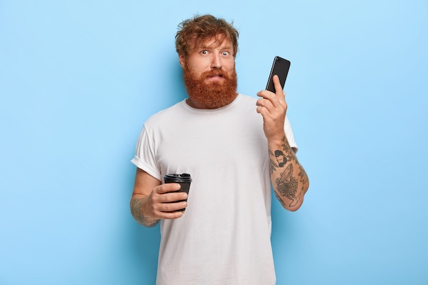 Zdezorientowany brodaty rudowłosy facet trzyma telefon komórkowy, odbiera telefon od nieznanej osoby, słyszy okropny głośny krzyk przez komórkę, pije kawę na wynos, nosi białą swobodną koszulkę
