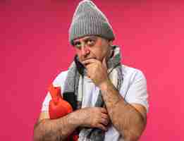 Bezpłatne zdjęcie zdezorientowany dorosły chory kaukaski mężczyzna z szalikiem na szyi w czapce zimowej kładzie rękę na brodzie i trzyma butelkę z gorącą wodą patrząc na bok odizolowany na różowej ścianie z miejscem na kopię