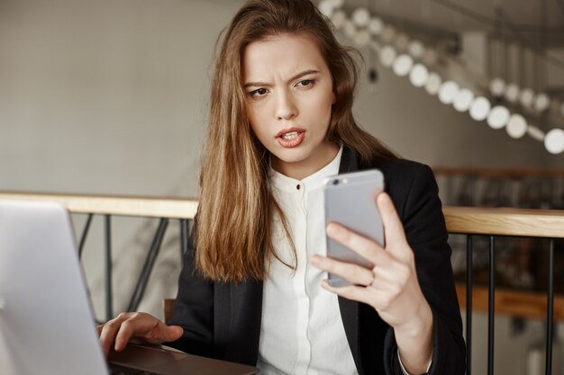 Zdezorientowany i niezadowolony bizneswoman patrząc na telefon komórkowy podczas pracy z laptopem w kawiarni