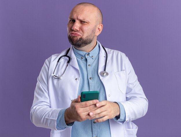 Zdezorientowany mężczyzna w średnim wieku, ubrany w szatę medyczną i stetoskop, trzymający telefon komórkowy z zaciśniętymi ustami odizolowanymi na fioletowej ścianie