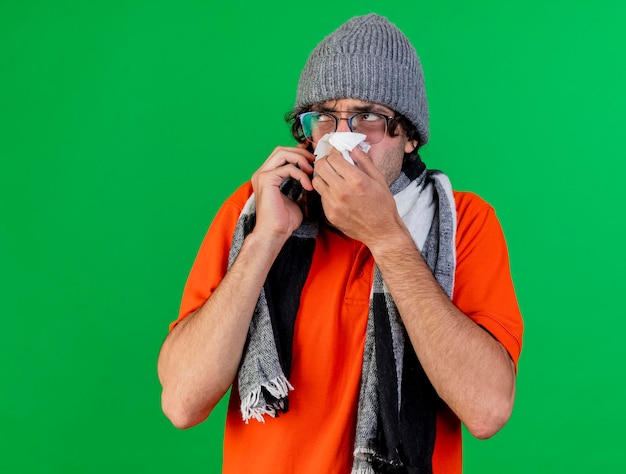 Bezpłatne zdjęcie zdezorientowany młody chory mężczyzna w okularach czapka zimowa i szalik rozmawia przez telefon wycierając nos patrząc w bok odizolowany na zielonej ścianie z miejscem na kopię