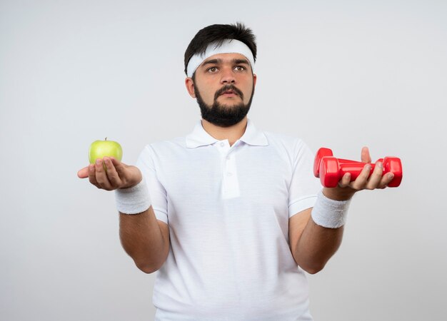Zdezorientowany młody człowiek sportowy patrząc z boku na sobie opaskę i opaskę trzymając hantle z jabłkiem na białym tle na białej ścianie