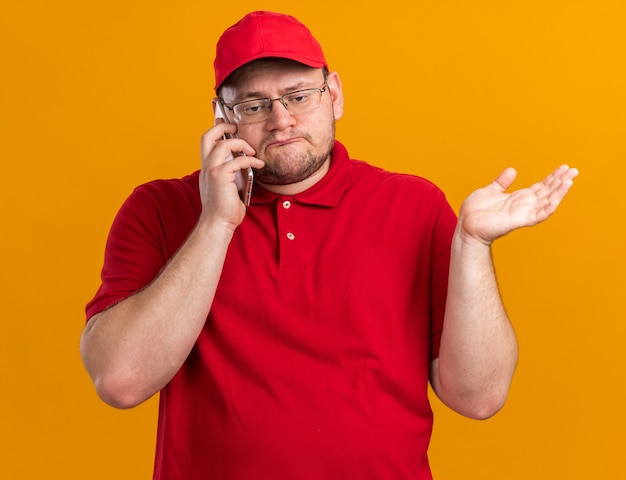 zdezorientowany młody dostawca z nadwagą w okularach optycznych rozmawia przez telefon odizolowany na pomarańczowej ścianie z kopią miejsca