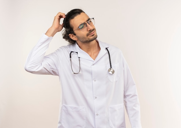 Zdezorientowany młody lekarz mężczyzna z okularami optycznymi na sobie białą szatę ze stetoskopem, kładąc rękę na głowie