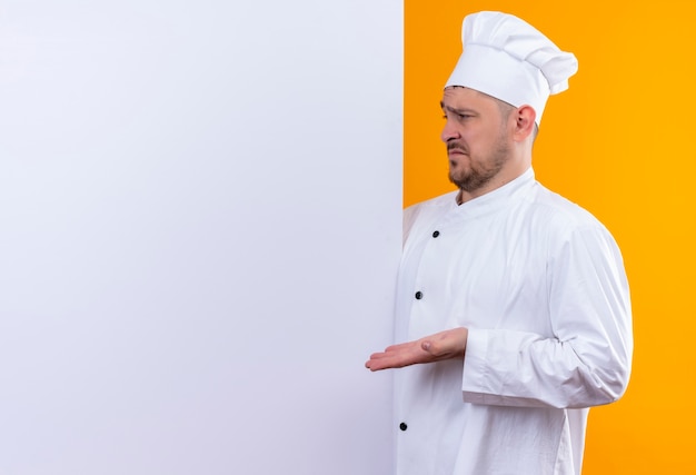 Zdezorientowany młody przystojny kucharz w mundurze szefa kuchni stojący za białą ścianą, wskazując ręką na to odizolowaną na pomarańczowej ścianie z miejscem na kopię