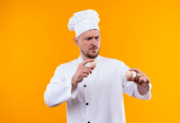 Zdezorientowany młody przystojny kucharz w mundurze szefa kuchni trzymający i patrzący na jajka izolowane na pomarańczowej ścianie
