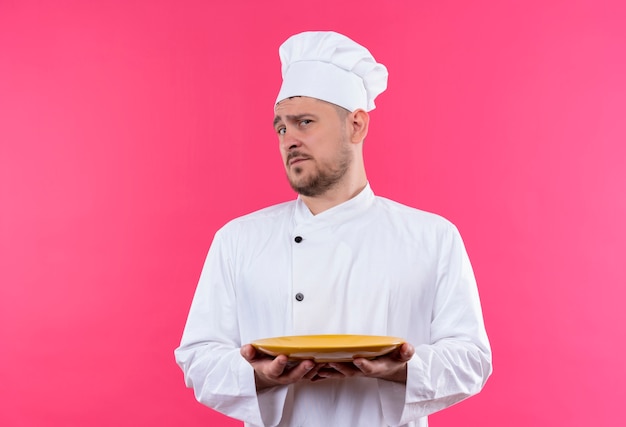 Zdezorientowany młody przystojny kucharz w mundurze szefa kuchni trzymający talerz odizolowany na różowej ścianie