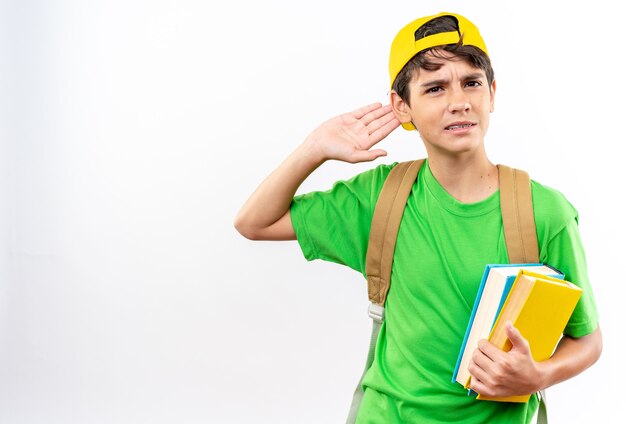 Zdezorientowany młody szkolny chłopiec noszący plecak z czapką trzymający książki pokazujące gest słuchania