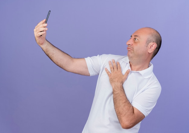 Zdezorientowany przypadkowy dojrzały biznesmen, trzymając rękę na klatce piersiowej i biorąc selfie na białym tle na fioletowym tle