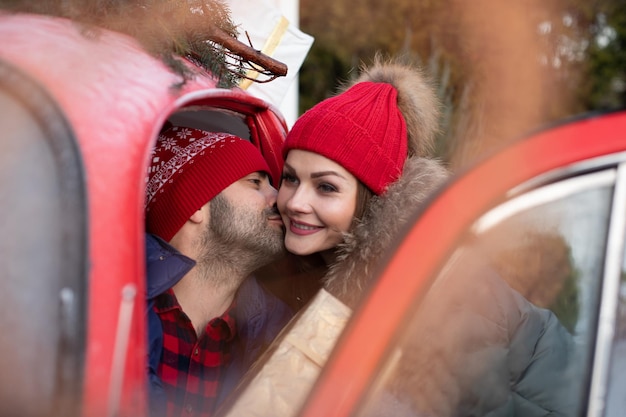 Bezpłatne zdjęcie zdjęcie atrakcyjnej kaukaskiej kobiety w ciepłych ubraniach niesie w samochodzie pudełka ze świątecznymi prezentami do swojego chłopaka i całuje go