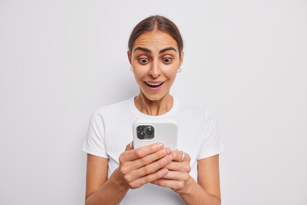 Zdjęcie brunetki czyta odebraną wiadomość z zapartym tchem, wpatrując się w ekran telefonu komórkowego, dostaje niesamowitą ofertę, która jest zachwycona, ma wyskakujące oczy, nosi casualową koszulkę odizolowaną na białej ścianie