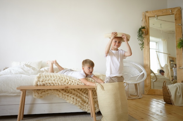 Zdjęcie dwóch uroczych kaukaskich uczniów, którzy bawią się w domu, grają razem w aktywne gry w sypialni rodziców, czują się szczęśliwi i beztroscy. Śliczne dzieci płci męskiej bawią się w domu