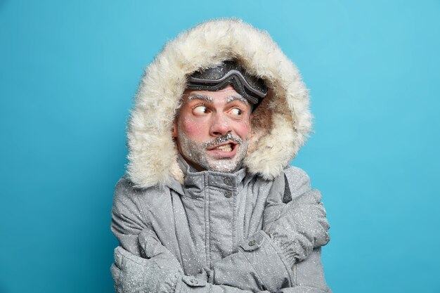 Zdjęcie Europejczyka drży z zimna po jeździe na deskorolce skrzyżowane ręce na ciele próbuje się ogrzać ubrany w szarą zimową kurtkę z futrzanym kapturem i rękawiczkami ma zamarzniętą twarz pokrytą lodem