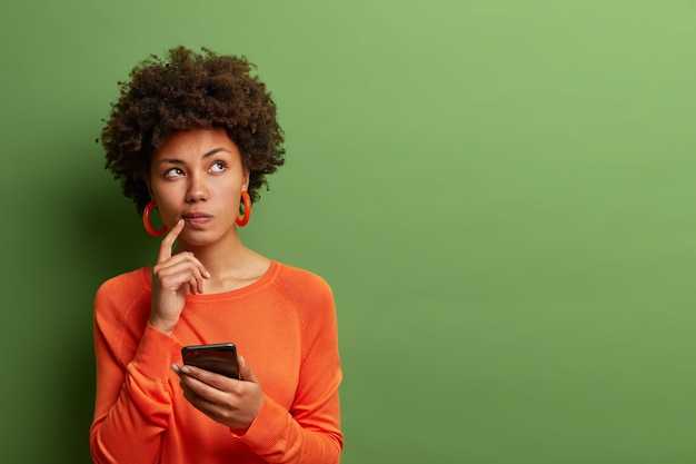 Zdjęcie ładnej etnicznej kobiety zastanawia się, jak odpowiedzieć na pytanie, głęboko się nad czymś zastanawia, używa nowoczesnego telefonu komórkowego, próbuje wymyślić dobry przekaz, trzyma palec wskazujący przy ustach, stoi w pomieszczeniu