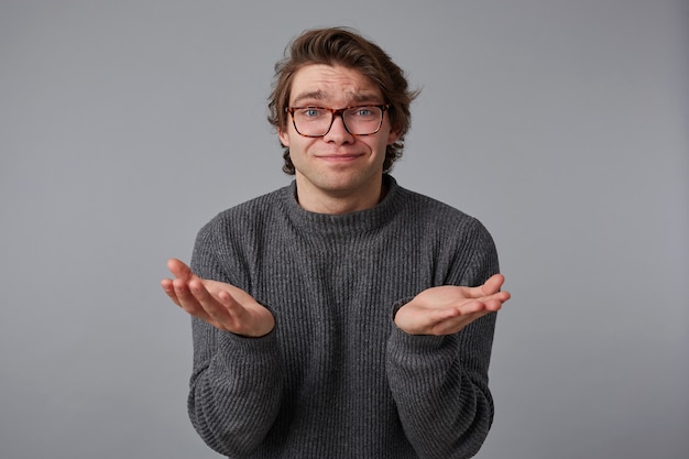 Zdjęcie młodego, przyjemnego mężczyzny w okularach, ubranego w szary sweter, stojącego na szarym tle i rozkładającego dłonie na bok.