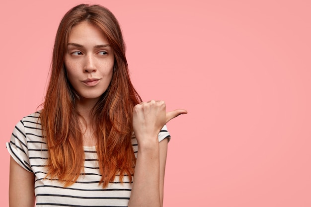 Bezpłatne zdjęcie zdjęcie młodej modelki z piegowatą zdrową skórą, wygląda tajemniczo na bok, wskazuje kciukiem na różową ścianę z miejscem na kopię