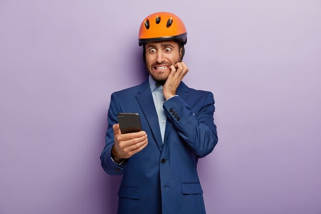 Zdjęcie nerwowego budowniczego gryzącego paznokcie ze wstydu, skupionego na smartfonie, czytającego zdziwione wiadomości z pracy, nosi formalny garnitur i kask