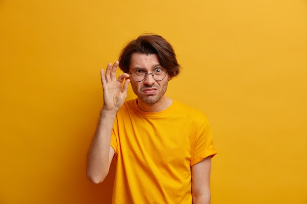Zdjęcie niewzruszonego niezadowolonego dorosłego mężczyzny kształtuje bardzo mały przedmiot, pokazuje coś małego, ma skrupulatny wygląd, nosi okrągłe przezroczyste okulary, odizolowane na żółtej ścianie