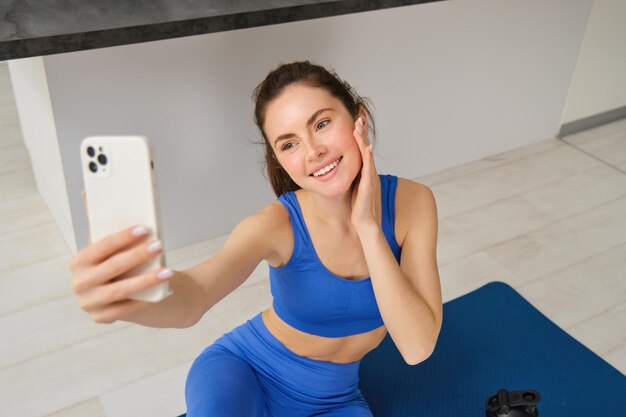 Zdjęcie pięknej wysportowanej dziewczyny siedzącej w domu na macie do jogi ze smartfonem robi selfie i trenuje