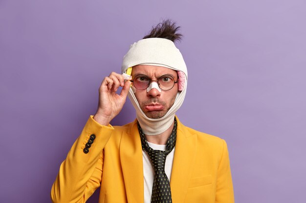 Zdjęcie ponurego niezadowolonego mężczyzny z siniakiem w okolicy oka, krwiakiem i wstrząsem mózgu, nosi bandaż, formalny garnitur i krawat, pobity przez okrutnych ludzi, odizolowane na fioletowej ścianie. Pojęcie problemu zdrowotnego