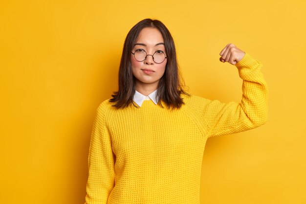 Zdjęcie poważnej brunetki Azjatki podnosi rękę i pokazuje, że jej siła ma silne mięśnie, stoi pewna siebie w domu, nosi żółty sweter i okrągłe okulary optyczne. Koncepcja mocy kobiet
