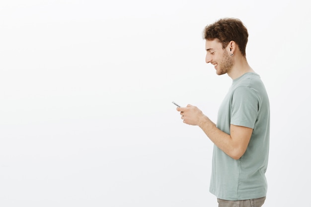 Zdjęcie profilowe przyjaznego przystojnego, młodego modela w kolczykach, trzymającego smartfona w dłoniach, szeroko uśmiechającego się do ekranu, zadowolonego z uroczej wiadomości od przyjaciela