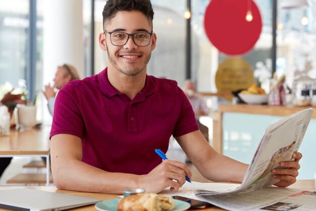 Zdjęcie przystojnego, zadowolonego faceta czyta najnowsze wiadomości w gazecie, zapisuje notatki w notatniku, nosi okulary i koszulkę, cieszy się pysznym rogalikiem. Ludzie i koncepcja pracy
