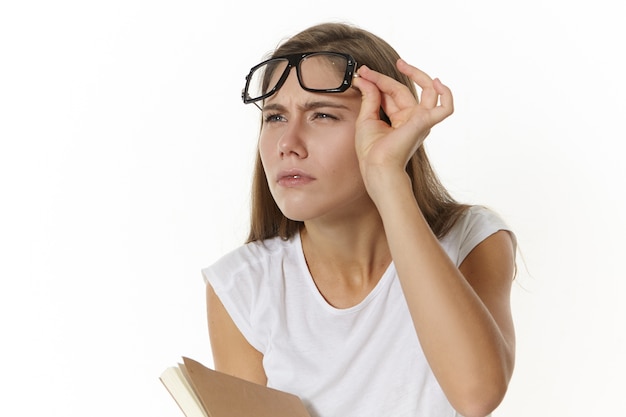 Bezpłatne zdjęcie zdjęcie skupionej, poważnej kaukaskiej nauczycielki z podręcznikiem, zdejmującej okulary i mrużących oczy, próbującej coś wyraźnie zobaczyć. student dziewczyna w okularach z pamiętnikiem