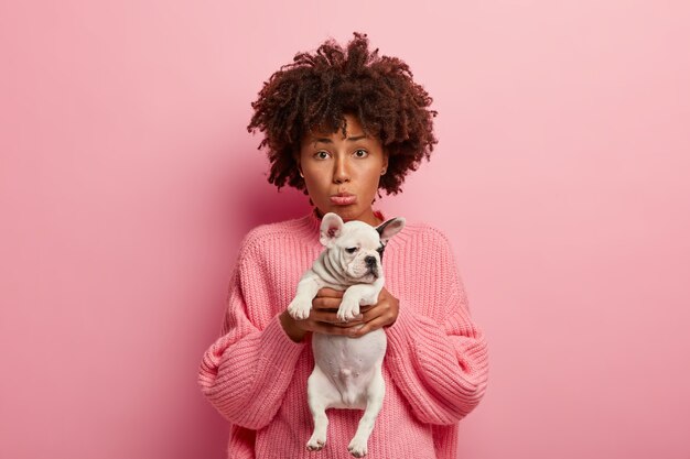 Zdjęcie smutnej, przygnębionej kobiety czuje się nieszczęśliwa, ponieważ jej zwierzak jest chory, nosi małego psa do weterynarza, zaciska usta, prosi o wyleczenie zwierzęcia, nosi luźny różowy sweter, pozuje razem ze szczeniakiem w pomieszczeniu na różowej ścianie
