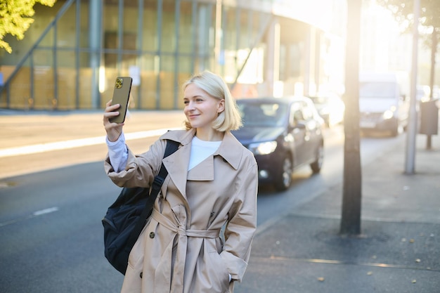 Zdjęcie stylowo uśmiechniętej blogerki robiącej selfie na ulicy, uśmiechającej się i pozującej dla mediów społecznościowych