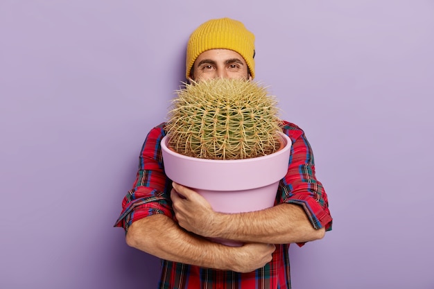 Bezpłatne zdjęcie zdjęcie szczęśliwego młodego męskiego hodowcy kwiatów obejmuje dużą doniczkę z kolczastym kaktusem, nosi stylowy kapelusz i kraciastą koszulę, ciesząc się, że otrzymuje w prezencie roślinę domową, odizolowane na fioletowej ścianie. koncepcja ogrodnictwa