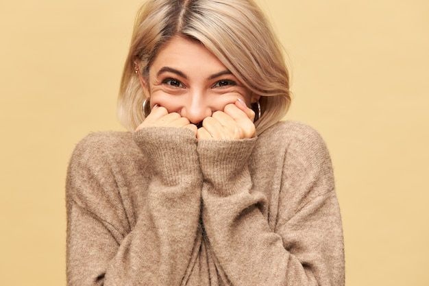 Zdjęcie szczęśliwej, radosnej młodej Europejki, która nie może ukryć swoich ekstatycznych emocji, jest w dobrym nastroju, przytłoczona pozytywnymi wiadomościami, chowa usta za dłońmi, nosi przytulny sweter z długimi rękawami