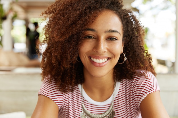Bezpłatne zdjęcie zdjęcie w głowę pięknej ciemnoskórej kobiety o radosnym wyrazie twarzy, fryzura afro, doskonale białe, równe zęby, zachwycony uśmiech. stylowa młoda kobieta african american spoczywa w pomieszczeniu