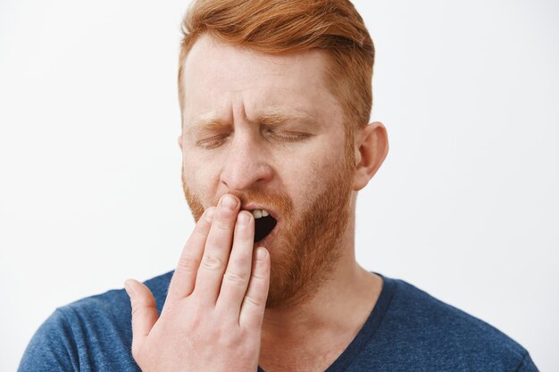 Zdjęcie w głowę zmęczonego atrakcyjnego przedsiębiorcy z rudymi włosami i brodą, ziewającego z zamkniętymi oczami, zakrywającego otwarte usta dłonią, zmęczonego, sennego po drzemce lub budzącego się wcześnie rano