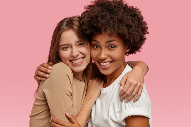 Zdjęcie wesołych, przyjaznych międzyrasowych kobiet obejmuje ciepłe uściski, radośnie się uśmiecha, pozuje do rodzinnego portretu, ubrane w luźne ubrania, odizolowane na różowej ścianie. Przyjaźń