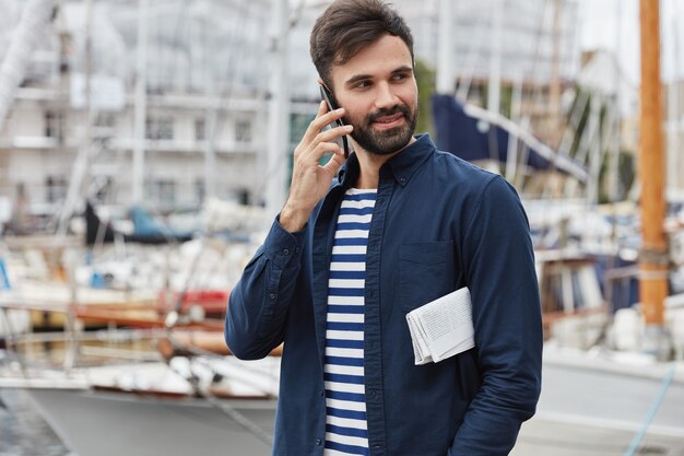 Zdjęcie zachwyconego hipstera z ciemną brodą w osłonie telefonu, spaceruje w pobliżu zatoki w porcie, nosi gazetę, patrzy na bok