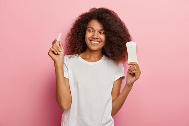 Zdjęcie zachwycony African American kobieta trzyma tampon i podpaski, ubrana w białą koszulkę, odizolowane na różowej ścianie. Kobiety, pms