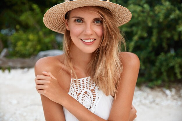 Zdjęcie zadowolonej atrakcyjnej kobiety o uroczym uśmiechu i cudownym wyglądzie, nosi stylowy słomkowy kapelusz, pozuje na zewnątrz na wybrzeżu, kąpie się w słońcu podczas upalnej letniej pogody. Koncepcja ludzie, sezon i odpoczynek