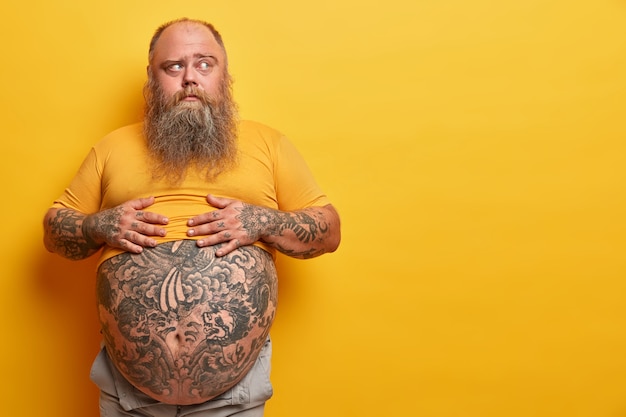 Zdjęcie zamyślonego mężczyzny z nadwagą trzyma ręce na dużym brzuchu z tatuażem, myśli i patrzy na bok, ma gęstą brodę, pozuje na żółtej ścianie. Otyły facet, który nie zdaje sobie sprawy, jak może wyglądać brzuch